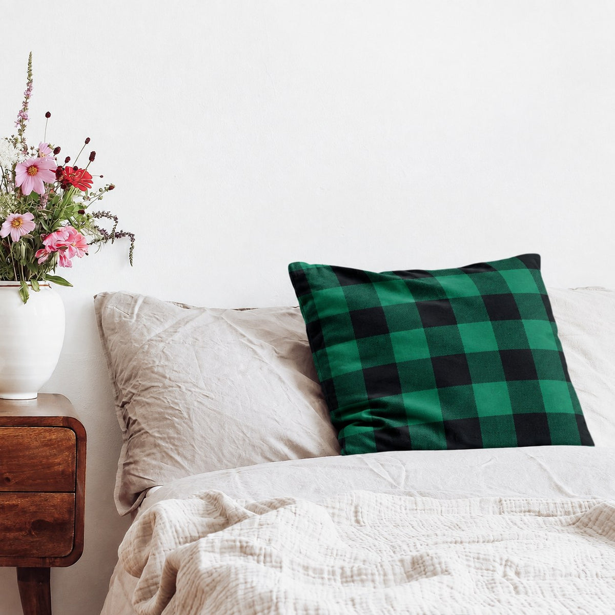 Coussin avec housse en flanelle à carreaux verts sur un lit, à côté d’une table de chevet avec des fleurs