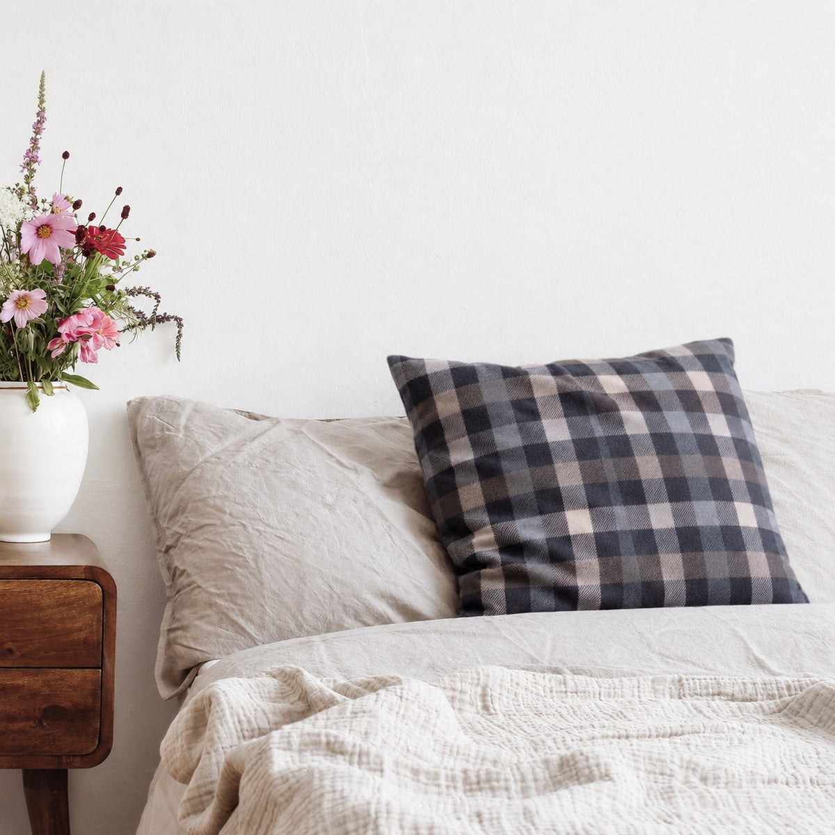 Coussin avec housse en flanelle à carreaux gris sur un lit, à côté d’une table de chevet avec des fleurs